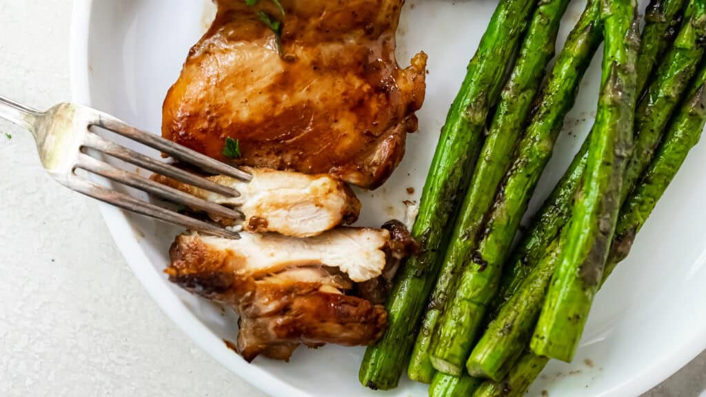 A plate with grilled chicken being cut with a fork next to a serving of cooked asparagus.