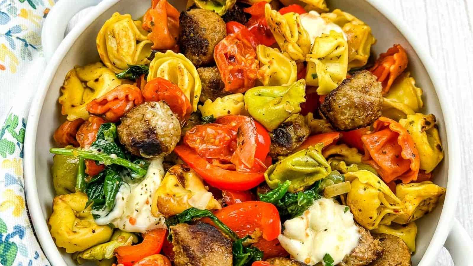 A close-up of a dish featuring colorful tortellini mixed with meatballs, spinach, and chopped red bell peppers, topped with melted cheese, served in a white bowl.