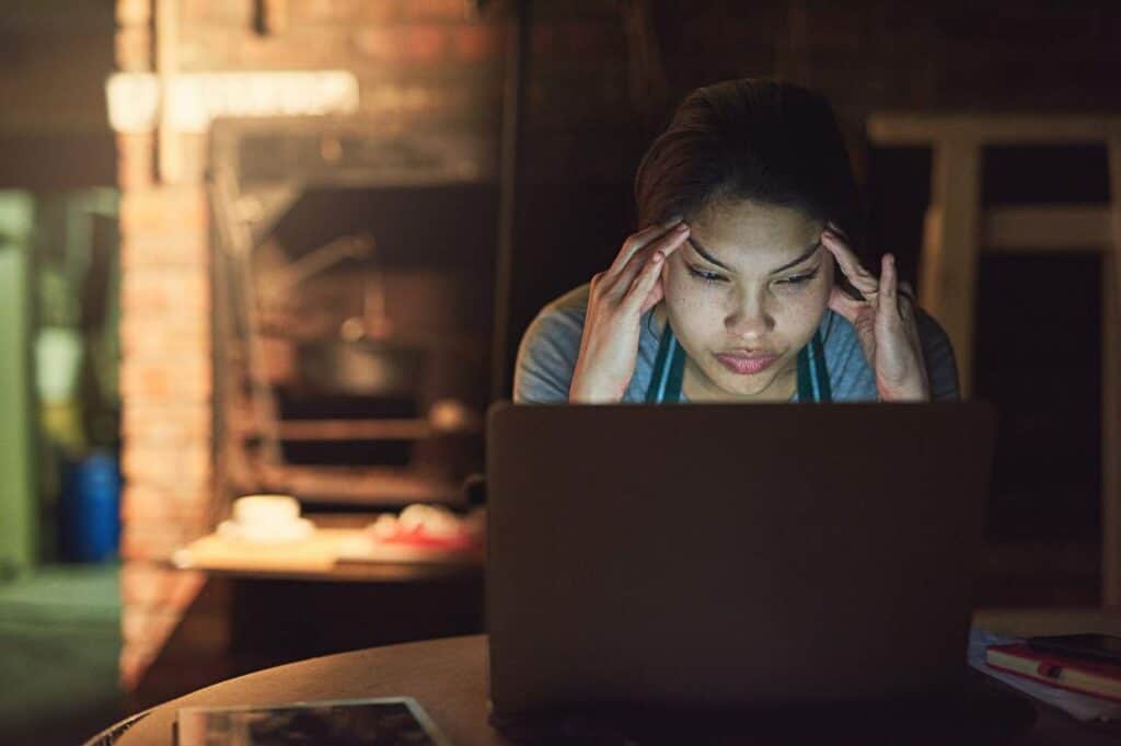 A person sitting at a table, looking at a laptop screen bathed in blue light with a focused, stressed expression, and holding their forehead with both hands.