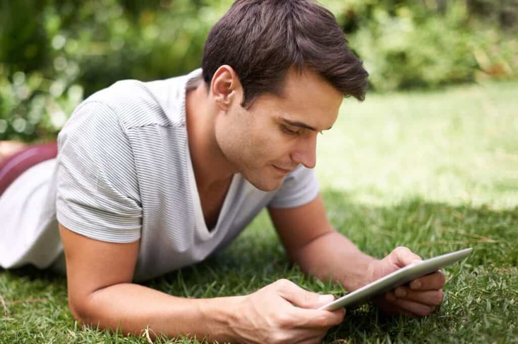 A man lies on the grass while looking at a tablet, likely protected against blue light. He is wearing a light-colored, striped t-shirt and has dark hair. The background is green and blurred, suggesting he is in a park or garden.