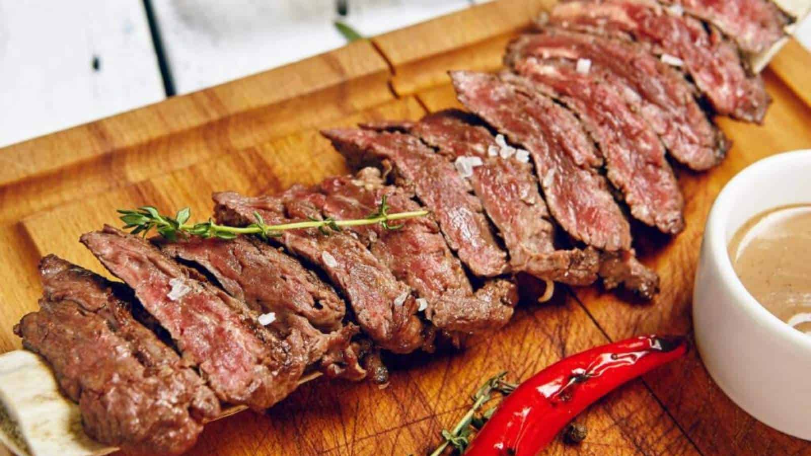 Sliced medium-rare grilled steak garnished with thyme and a red chili pepper on a wooden cutting board.