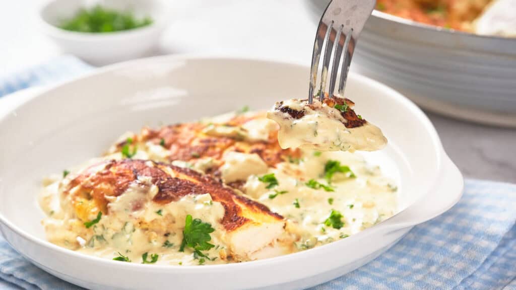 A fork lifting a bite of creamy, herb-covered chicken breast from a white bowl.