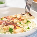 A fork lifting a bite of creamy, herb-covered chicken breast from a white bowl.