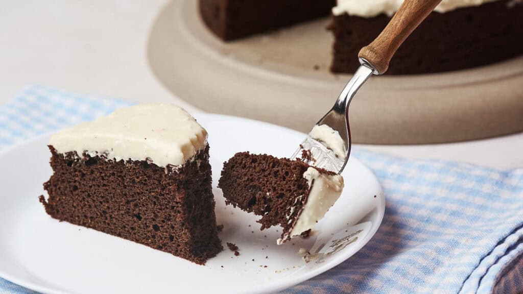 A slice of chocolate cake with white frosting on a white plate, being cut with a fork.