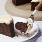 A slice of chocolate cake with white frosting on a white plate, being cut with a fork.