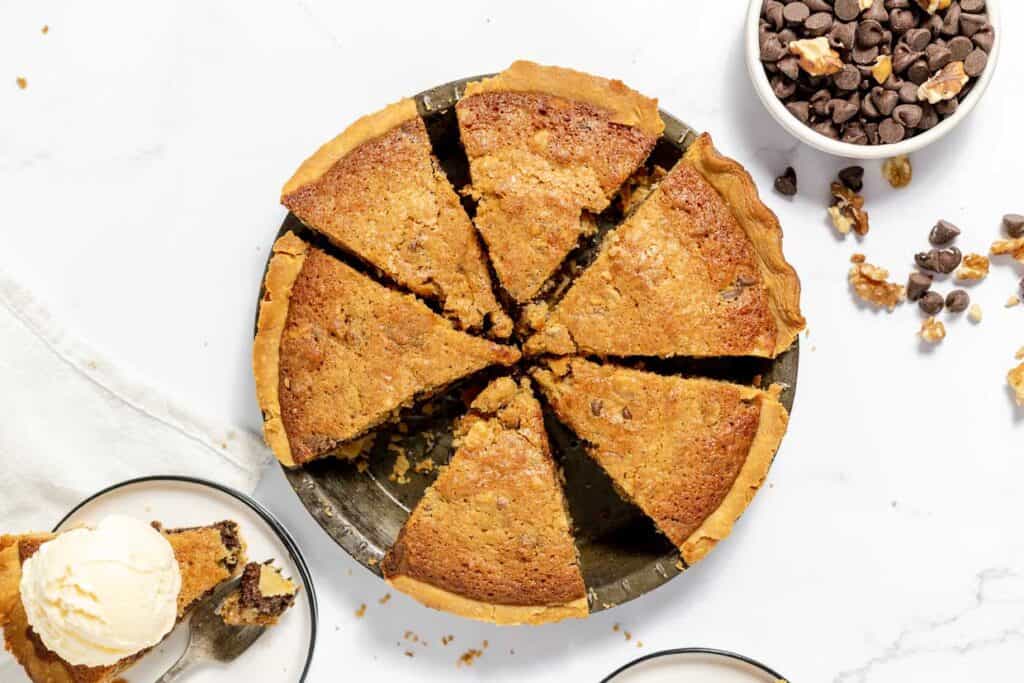 A round pie cut into eight slices on a pie dish, accompanied by a bowl of chocolate chips and a smaller bowl of walnut pieces. A plate with a slice of pie topped with ice cream is nearby.