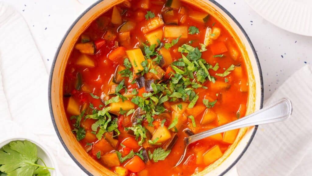 A pot of Italian vegetable stew with a spoon, garnished with chopped green herbs, on a white tablecloth with a small bowl of parsley on the side.