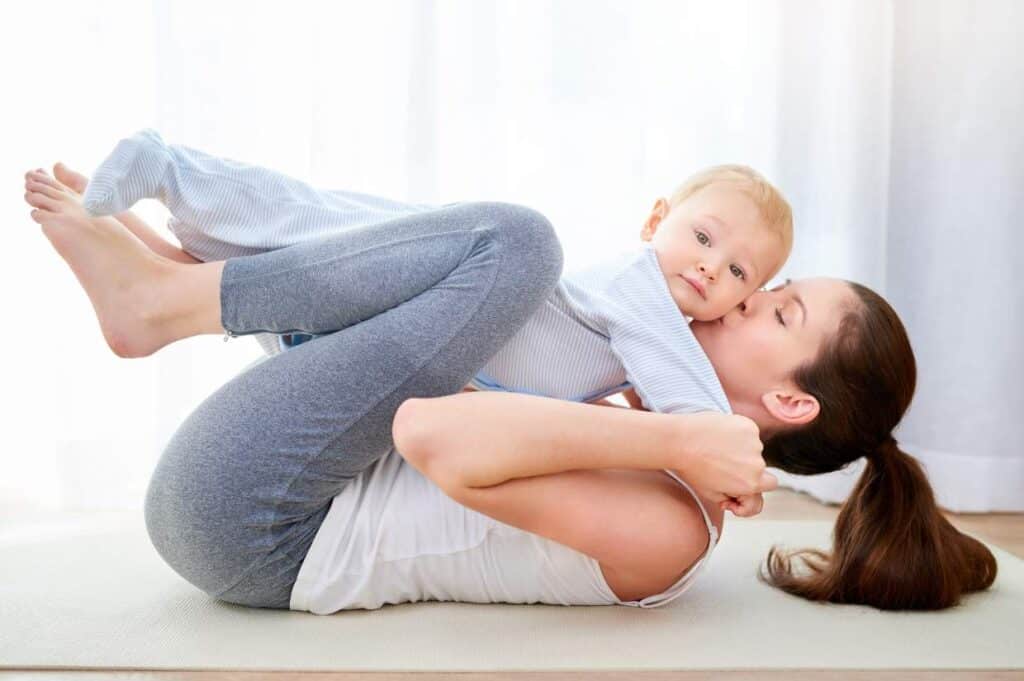 A woman lies on her back on a mat, lifting a baby who rests on her legs.
