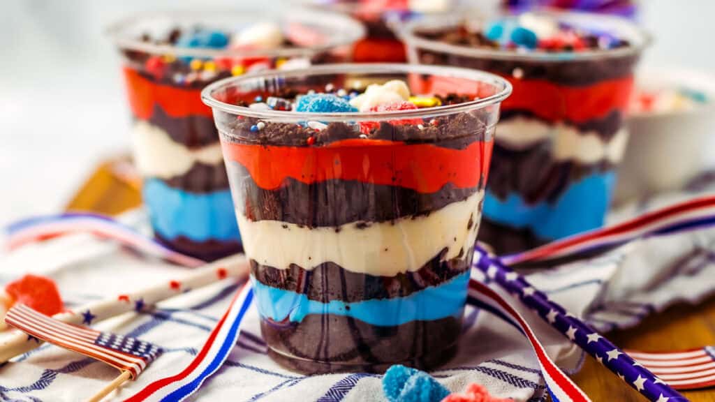Three patriotic-themed layered desserts in clear cups, featuring red, white, and blue layers, accented with sprinkles and decorations. American flag toothpicks are nearby on a festive cloth.