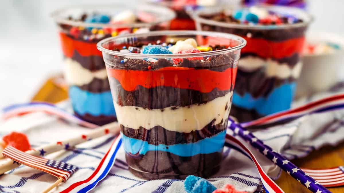 Layered patriotic desserts in clear cups featuring red, white, and blue layers of cream and chocolate, adorned with festive toppings, placed on a cloth with decorative red, white, and blue accents.