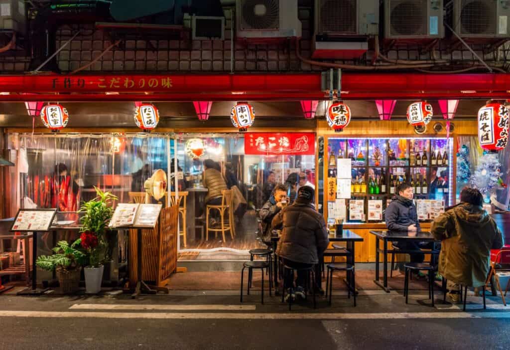 People dine at an outdoor and indoor restaurant with red lanterns and heaters in a vibrant setting. The establishment features colorful signage and a variety of beverages displayed on shelves.