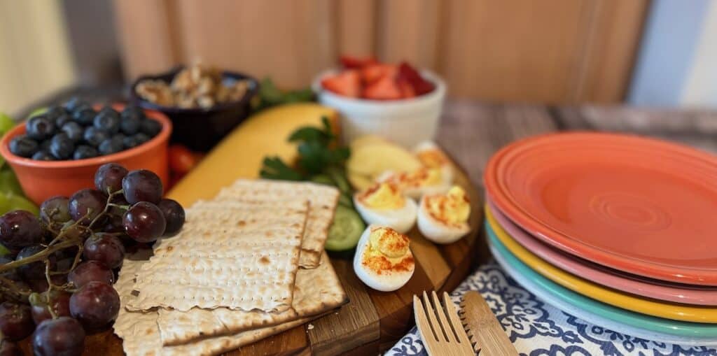 A wooden board with crackers, grapes, blueberries, walnut pieces, sliced cheese, and deviled eggs. Nearby are bowls of strawberries and a stack of colorful plates with wooden cutlery.