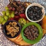 A wooden platter with grapes, walnuts, chocolate-covered almonds, kiwi slices, dried apricots, blueberries, strawberries, and apple slices on a patterned tablecloth.