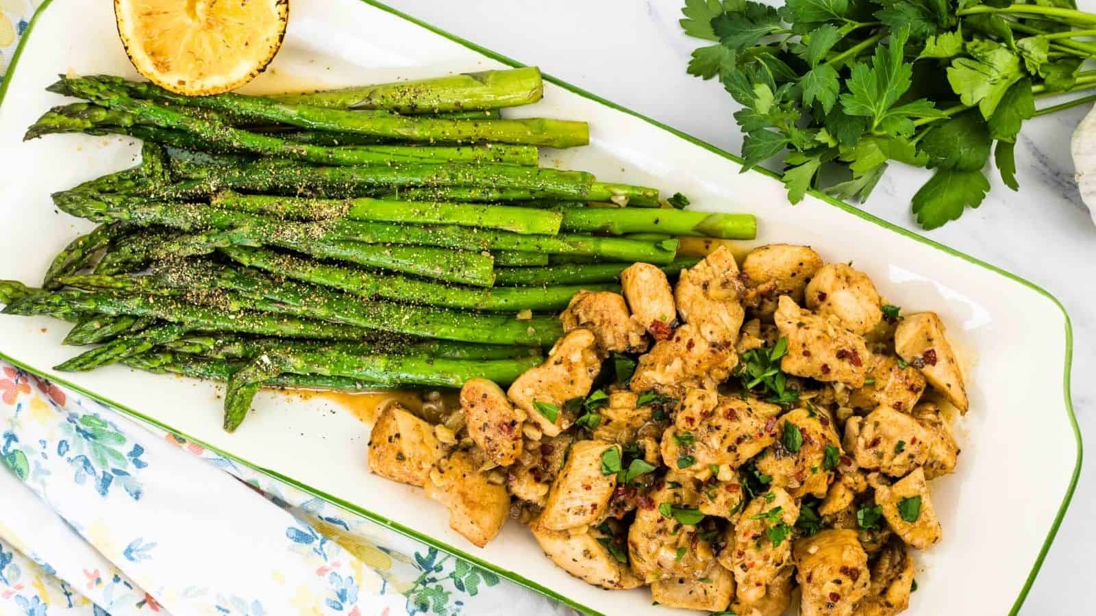A plate with seasoned asparagus and diced chicken topped with herbs. A halved grilled lemon and fresh parsley are nearby on a white floral tablecloth.