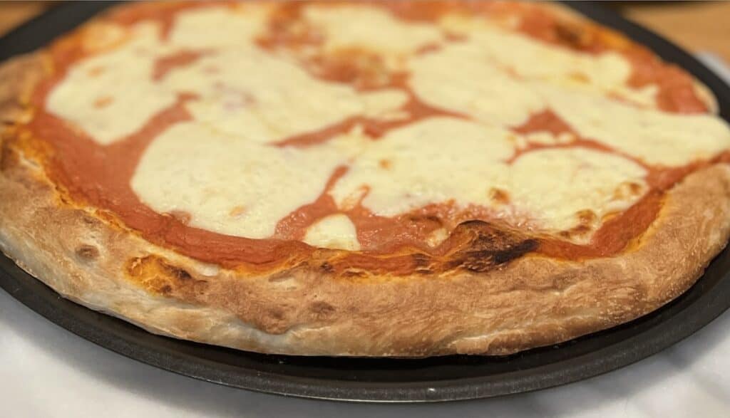 A close-up of a freshly baked pizza with a thick crust, tomato sauce, and melted cheese. The pizza is on a round metal tray.