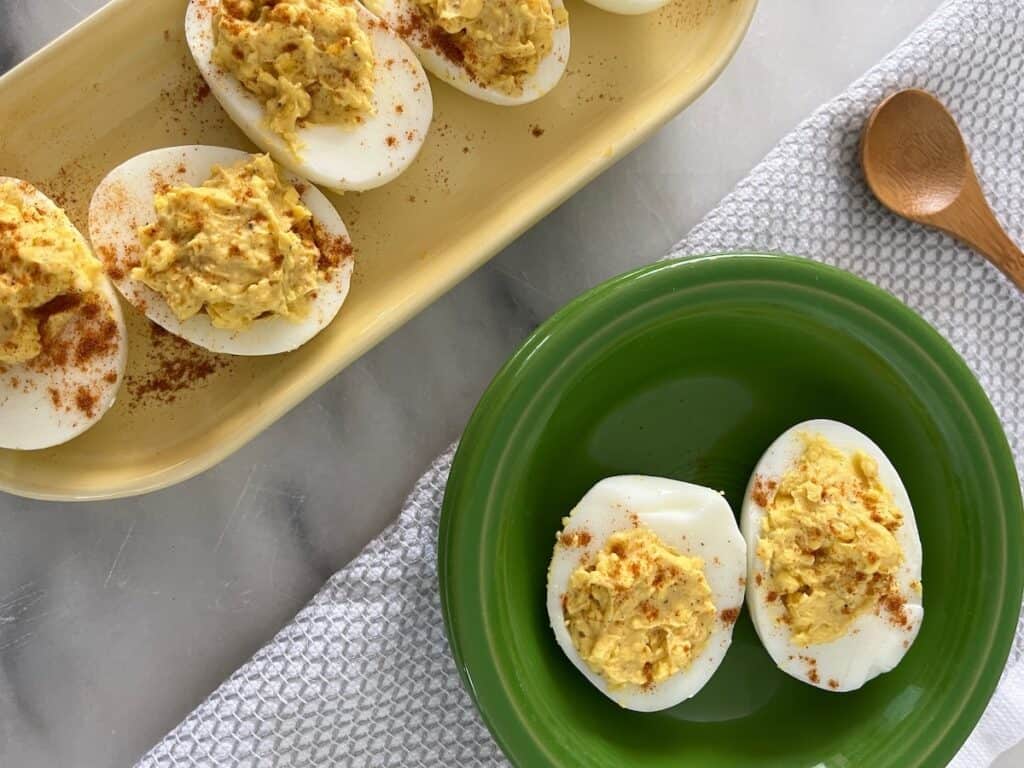 Two plates of deviled eggs garnished with paprika. One plate is green with two eggs, and the other is yellow with several eggs. A wooden spoon and a textured cloth are visible nearby.