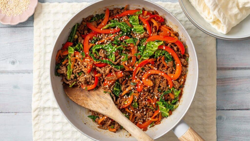 A skillet filled with a colorful stir-fry of ground meat, red bell peppers, leafy greens, and sesame seeds, placed on a light-colored cloth with a wooden spoon. Plates and wraps are nearby.