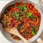 A skillet filled with a colorful stir-fry of ground meat, red bell peppers, leafy greens, and sesame seeds, placed on a light-colored cloth with a wooden spoon. Plates and wraps are nearby.