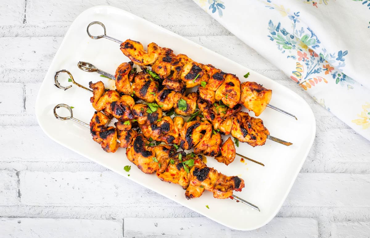 A rectangular white plate with four Sweet & Spicy Grilled Harissa-Honey Chicken Skewers garnished with chopped herbs.
