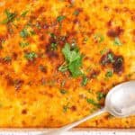 A baked casserole with a browned cheese crust topped with chopped parsley, served in a rectangular dish with a spoon on the side.