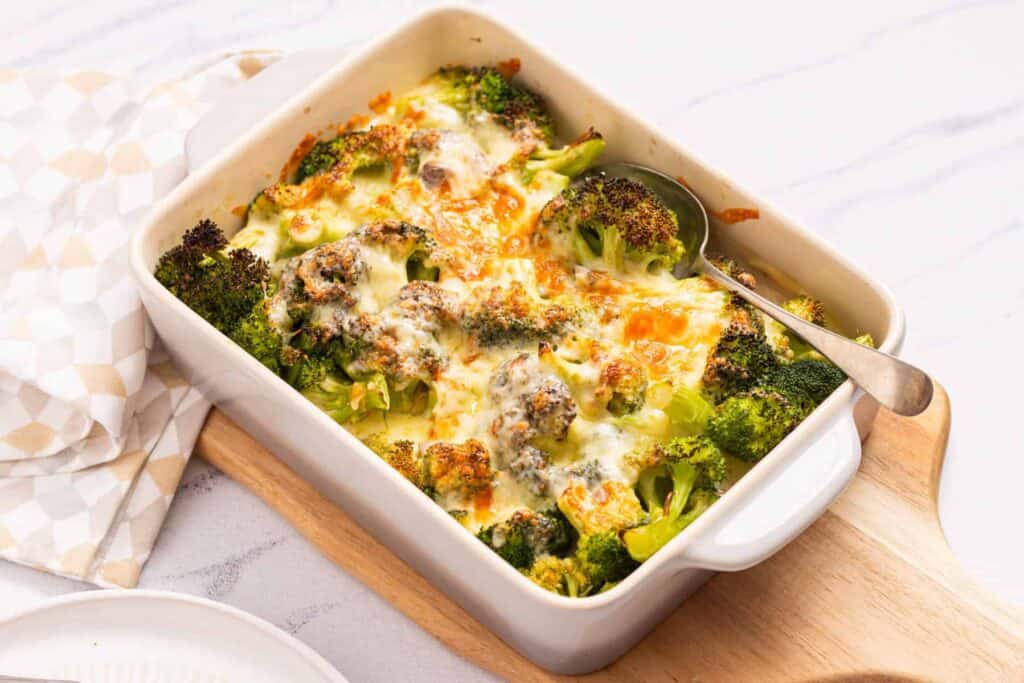 A baked broccoli casserole with melted cheese in a white dish, placed on a wooden board with a spoon inside. A napkin and a partial view of a plate are near the warm, inviting casseroles.