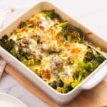 A baked broccoli casserole with melted cheese in a white dish, placed on a wooden board with a spoon inside. A napkin and a partial view of a plate are near the warm, inviting casseroles.