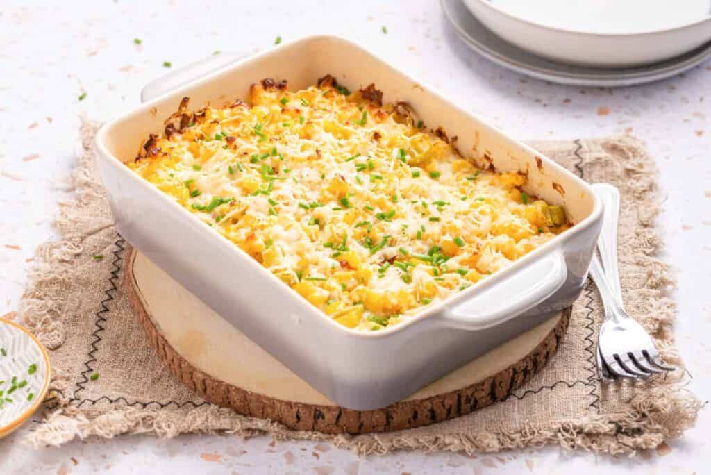 A baked casserole in a rectangular dish, one of many delightful casseroles, is garnished with chopped herbs and placed on a wooden serving board with a fork beside it. Plates are visible in the background.