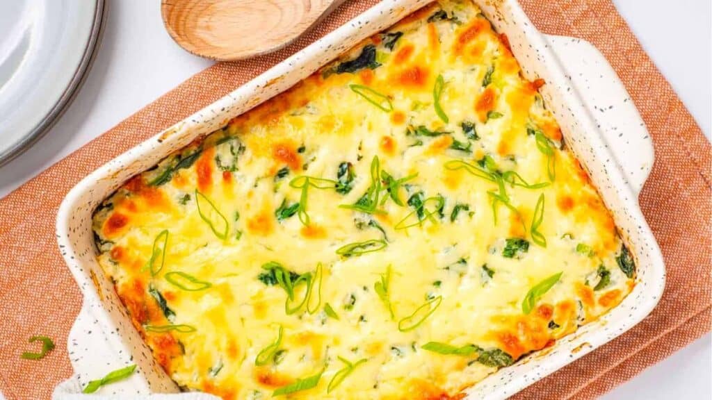 A baked casserole dish containing a cheesy, green herb-topped meal rests on an orange mat. A wooden spoon lies beside the casseroles dish.