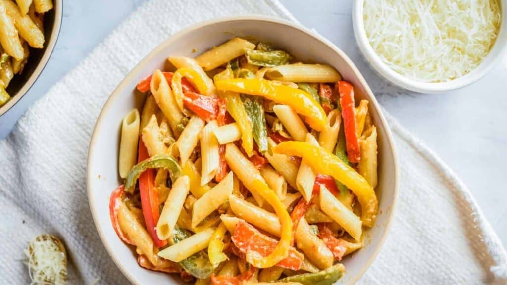 A bowl of cooked penne pasta mixed with colorful bell peppers, placed on a white cloth beside a small bowl of grated cheese.
