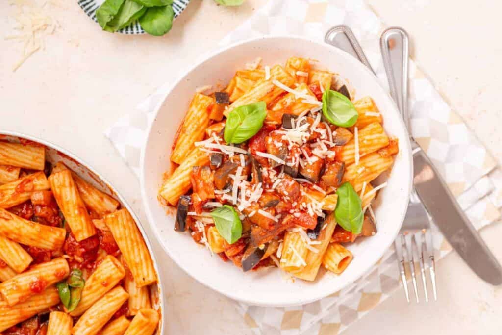 A bowl of rigatoni pasta with tomato sauce, eggplant, grated cheese, and basil leaves, next to a pan with more pasta and a fork and knife set.