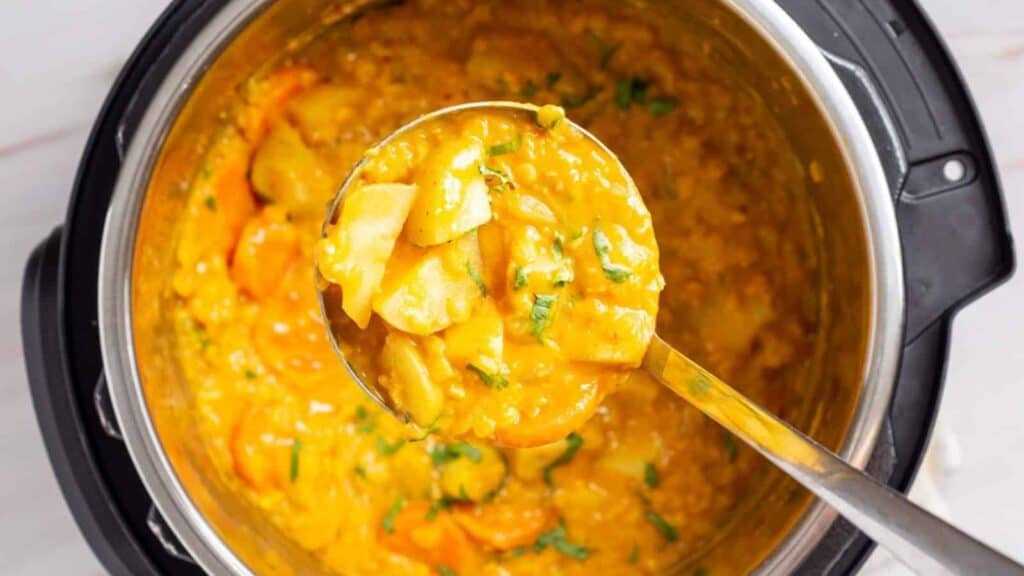 Ladle scooping a portion of thick, yellow lentil and vegetable curry from an instant pot. The curry contains pieces of potato, carrot, and greens.