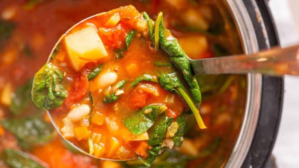 A ladle holds vegetable soup with spinach, tomatoes, beans, and diced vegetables over a bowl filled with the soup.