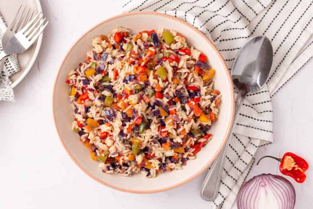 A bowl of colorful rice mixed with diced vegetables is placed on a white and black-striped napkin. A fork and spoon are on the side, with a sliced red onion and a small red pepper nearby.