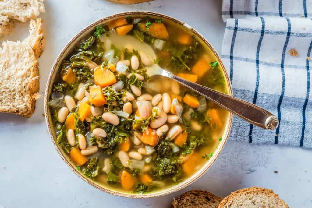 A bowl of vegetable soup containing white beans, kale, and diced carrots, with a spoon inside. Slices of bread and a checkered cloth napkin are placed beside the bowl.