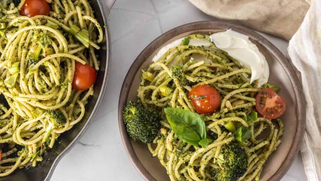 A plate of spaghetti pasta with broccoli, cherry tomatoes, and a green pesto sauce, garnished with fresh basil leaves, next to a skillet containing more of the same pasta dish.