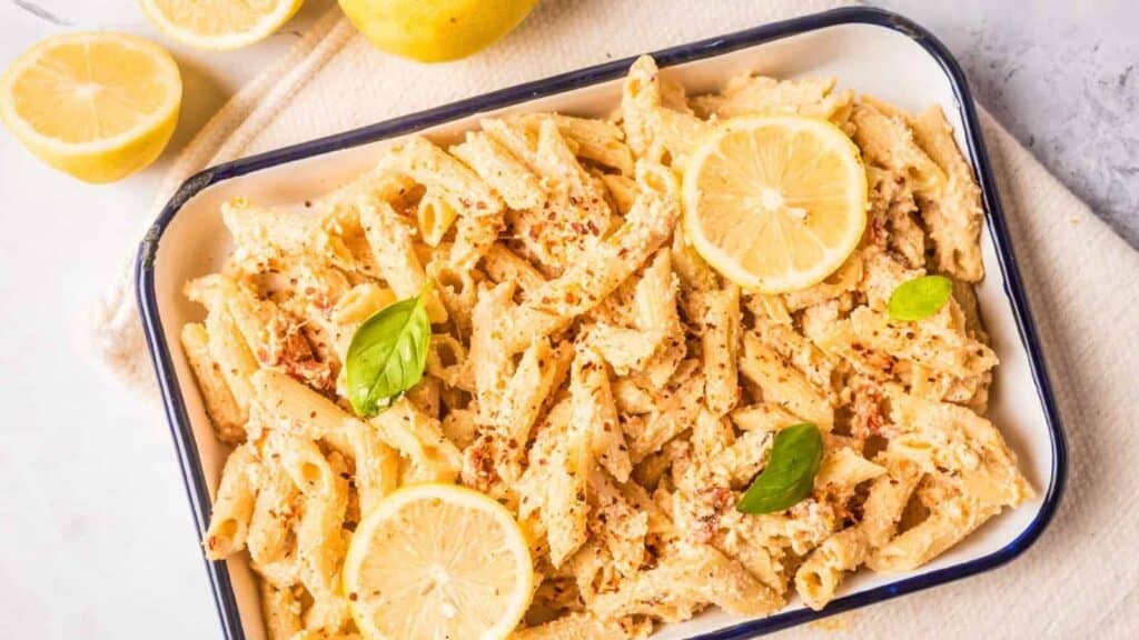 A rectangular white dish contains baked penne pasta with creamy sauce, lemon slices, basil leaves, and a sprinkle of seasoning. Lemons are placed nearby on a light surface.