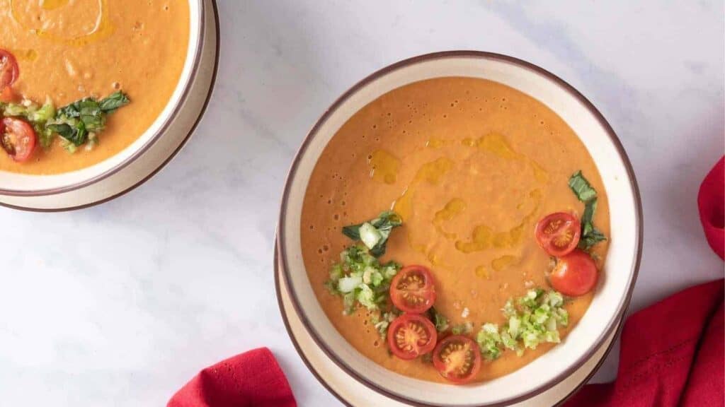 Two bowls of gazpacho garnished with chopped vegetables and halved cherry tomatoes, placed on a light-colored surface with a red cloth nearby.