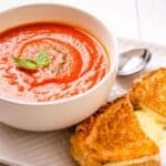 A bowl of tomato soup with a basil garnish next to a grilled cheese sandwich on a white plate with a spoon.