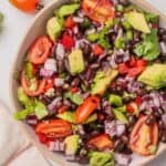 A bowl filled with a colorful salad containing cherry tomatoes, avocado, black beans, red onions, cilantro, and bell peppers. A white and red striped cloth and a lime are nearby.