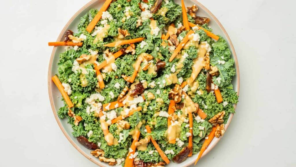 A plate of kale salad topped with chopped carrots, walnuts, feta cheese, and a light dressing.