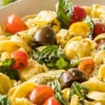 A close-up of a pasta salad featuring tortellini, halved cherry tomatoes, spinach, and other greens, all mixed and garnished with herbs and grated cheese.