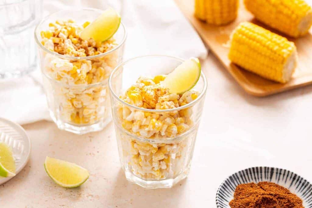 Two glasses of corn salad topped with lime wedges sit on a table, with more corn on a cutting board and a bowl of spices nearby.