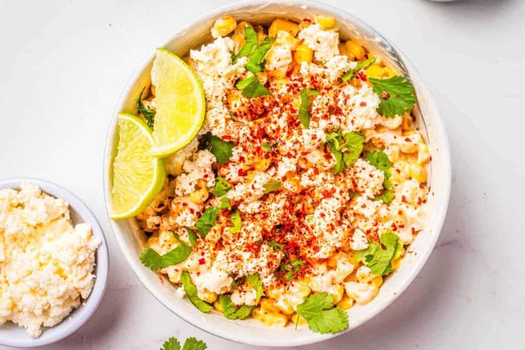 A bowl of Mexican street corn topped with crumbled cheese, paprika, chopped cilantro, and lime wedges on the side, with a small bowl of extra cheese nearby.