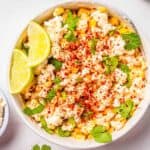 A bowl of Mexican street corn topped with crumbled cheese, paprika, chopped cilantro, and lime wedges on the side, with a small bowl of extra cheese nearby.