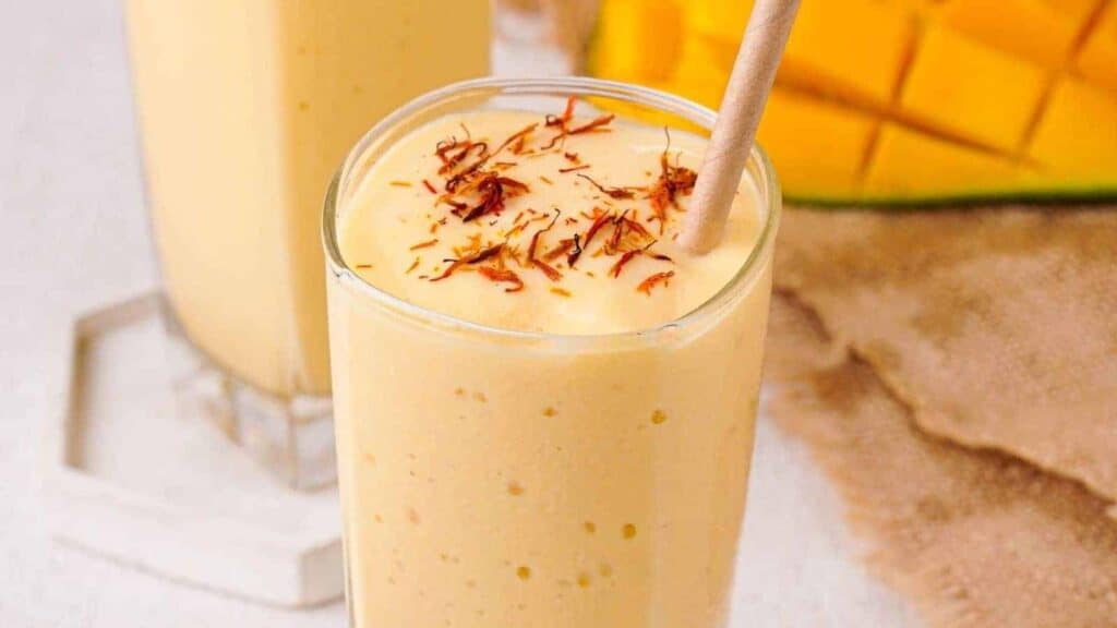 A glass of mango lassi topped with saffron strands, with diced mangoes and another glass of lassi in the background.