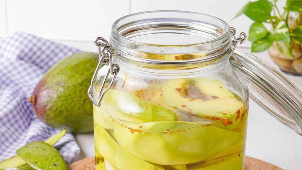 A glass jar filled with sliced pickled avocados, with an open lid. A whole mango and a partially peeled mango are placed next to the jar. A checkered cloth is in the background.