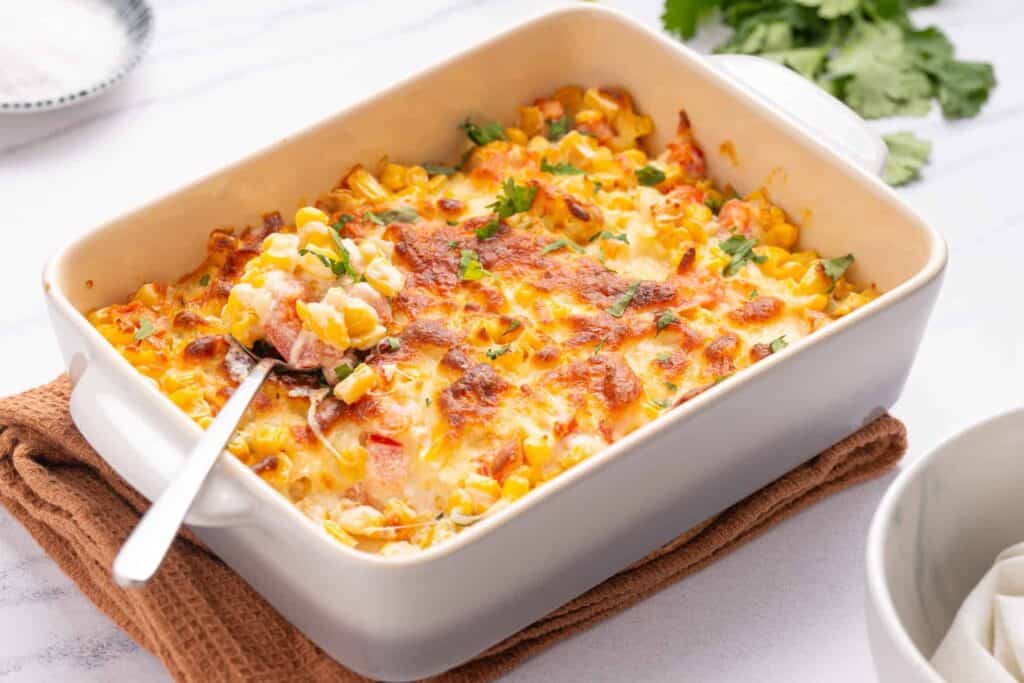 A baked corn dip with a golden, cheesy top in a white rectangular dish, garnished with chopped herbs, and a spoon inserted in the dish, placed on a brown cloth.