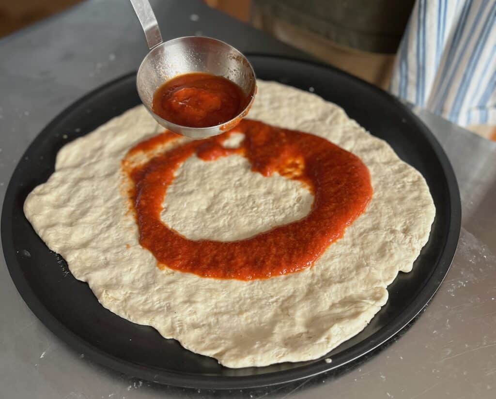 A ladle pours tomato sauce onto an uncooked pizza dough placed on a round black pan.