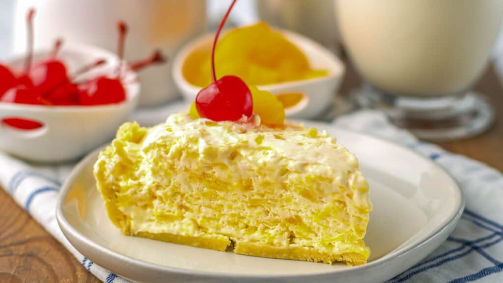 A slice of chilled pineapple pie topped with a cherry, on a white plate. Surrounding it are additional cherries, pineapple pieces, and a beverage in the background.
