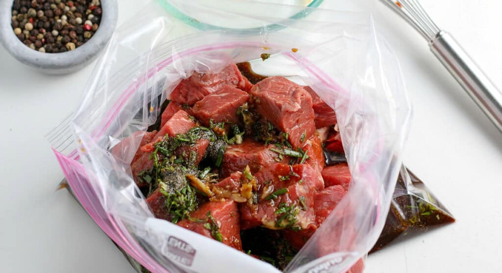 Chunks of raw beef marinating in a plastic ziplock bag with herbs and spices, placed on a white surface near a bowl of peppercorns and a whisk.